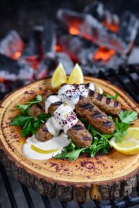 كفتة لحم غنم كيتو مشوية على الفحم مع صوص طحينة كريمي، بقدونس طازج، سماق، وشرائح ليمون على لوح خشبي ريفي