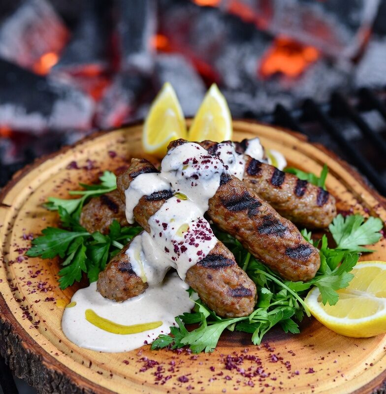 كفتة لحم غنم كيتو مشوية على الفحم مع صوص طحينة كريمي، بقدونس طازج، سماق، وشرائح ليمون على لوح خشبي ريفي